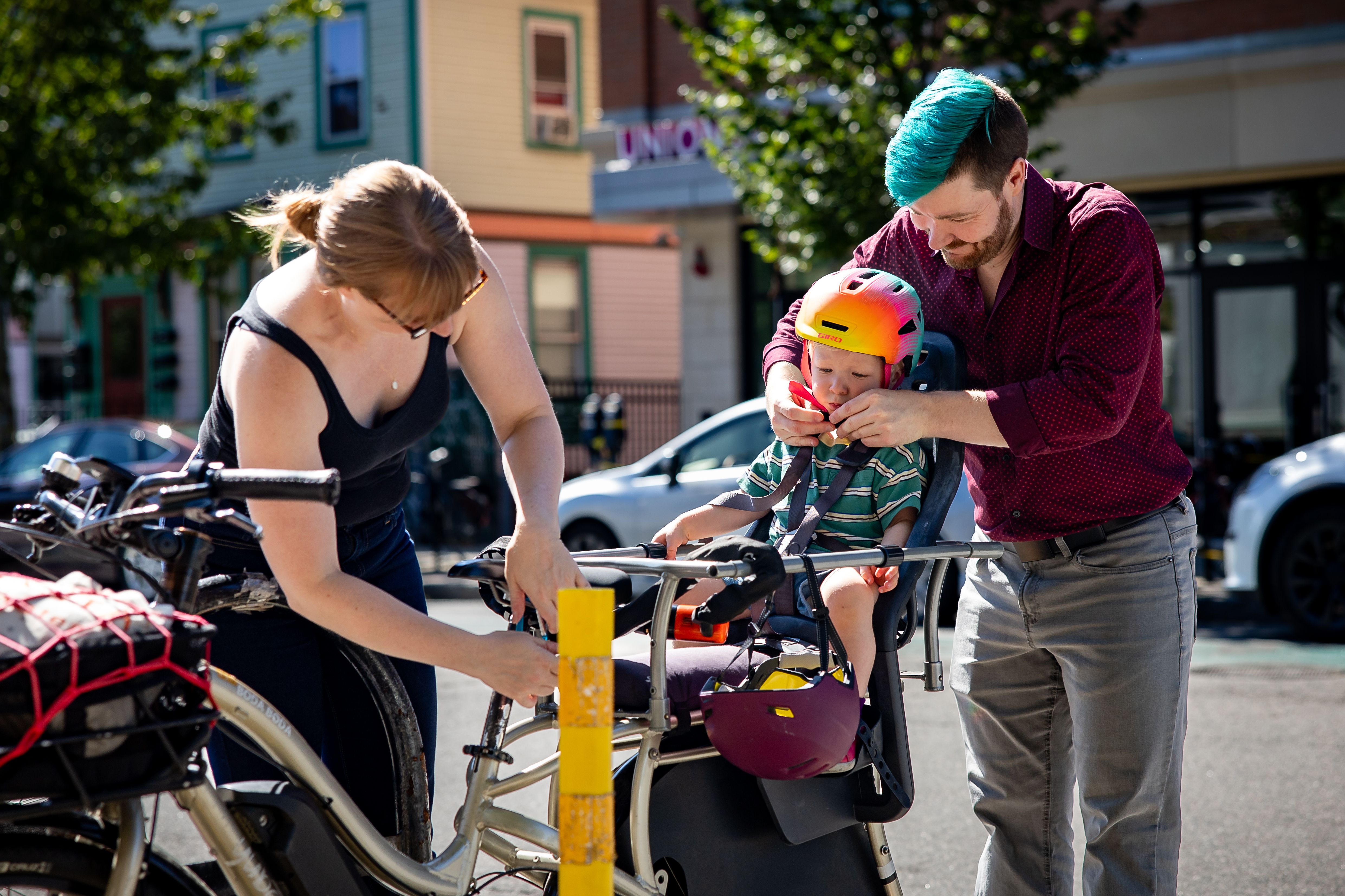 Derrick strapping his son into a bike toddler seat, while his wife unlocks it from the bike rack.